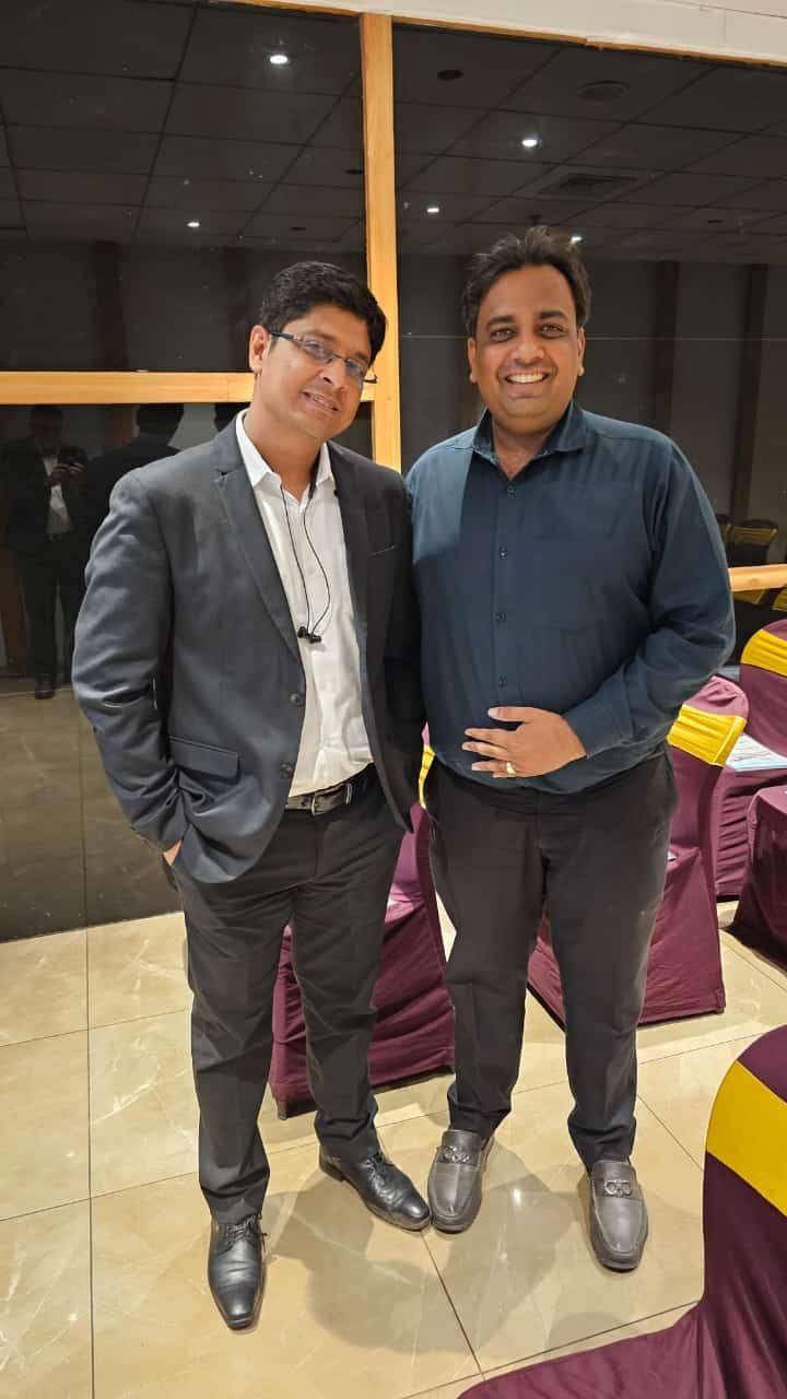 Two men smiling and posing indoors, both wearing formal suits.